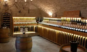 Cave voûtée du **domaine de Montmain** avec étagères en bois illuminées présentant de nombreuses bouteilles de vin, tonneaux et lustre ancien.