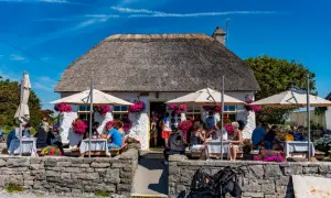 Café pittoresque avec toit de chaume, parasols et fleurs. Des gens dégustent un repas en plein air, peut-être un délicieux aran poisson.