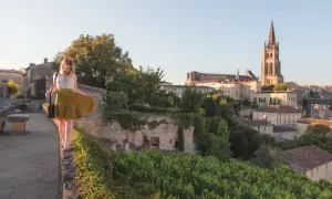 Une femme en jupe jaune marche sur un muret, surplombant le village de Saint-Émilion. La route des vins s'étend à perte de vue.