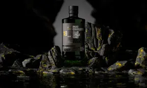 Bouteille de whisky Port Charlotte 18 ans d'âge, produit par la distillerie Bruichladdich, posée sur des rochers sombres aux reflets dorés.