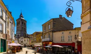 Une rue animée de la ville de Cognac, avec une tour d'église imposante en arrière-plan. Des terrasses de cafés invitent à la détente sous le soleil.