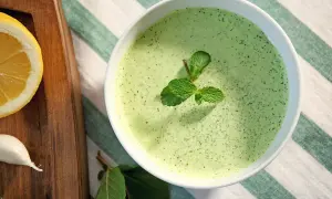 Un bol de sauce à la menthe fraîchement préparée, garni de feuilles de menthe. À côté, une tranche de citron et une gousse d'ail sur une planche en bois.