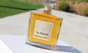Bouteille de "Le Baron Grande Réserve Gérard", un spiritueux ambré, posée sur un socle blanc, avec un arrière-plan flou évoquant un jardin et un chemin, rappelant l'élégance d'un chateau vaudois.