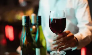 Main d'un homme tenant un verre de vin rouge, prêt pour une dégustation de vin Beaujolais. Des bouteilles floues en arrière-plan.