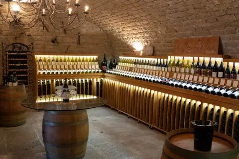 Cave voûtée du **domaine de Montmain** avec étagères en bois illuminées présentant de nombreuses bouteilles de vin, tonneaux et lustre ancien.