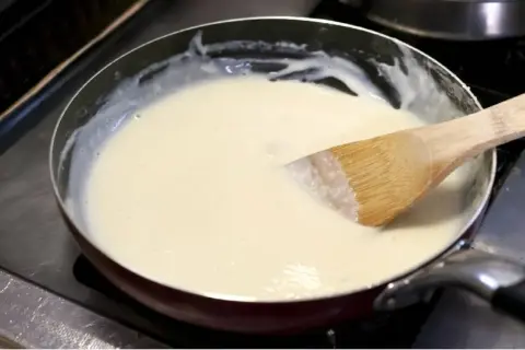 Une spatule en bois remue une sauce onctueuse dans une poêle. La préparation crémeuse, une sauce vin blanc et crème, est prête à être servie.