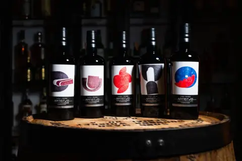 Cinq bouteilles de blend whisky Artist-13, ornées d'étiquettes artistiques colorées, reposent sur un fût de whisky.