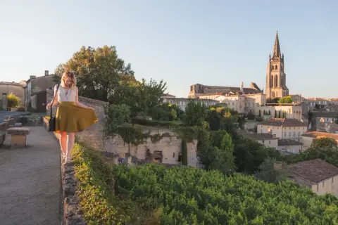Une femme en jupe jaune marche sur un muret, surplombant le village de Saint-Émilion. La route des vins s'étend à perte de vue.
