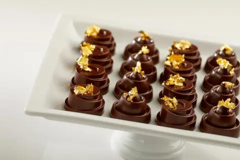 Douze chocolats fins, une composition alimentaire luxueuse, ornés de feuilles d'or comestibles, présentés sur un plat blanc.