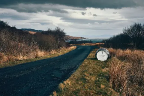 Route sinueuse menant à la distillerie Bunnahabhain, avec un fût indiquant la direction. Paysage écossais sauvage sous un ciel menaçant.