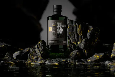 Bouteille de whisky Port Charlotte 18 ans d'âge, produit par la distillerie Bruichladdich, posée sur des rochers sombres aux reflets dorés.