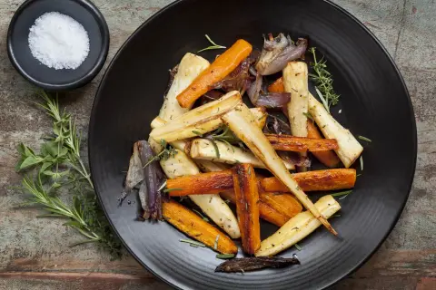 Légumes rôtis, carottes et panais, parsemés d'herbes. Un navet doré, tendre et savoureux, se mêle aux autres légumes sur l'assiette noire.