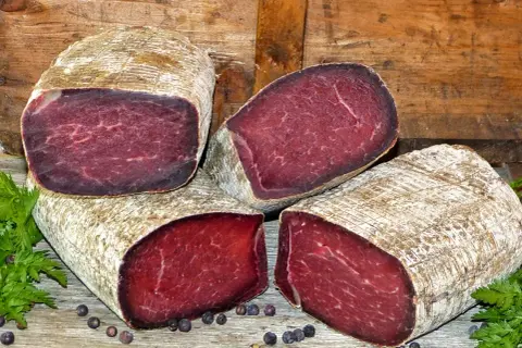 Quatre morceaux de viande des Grisons, d'origine suisse, sont présentés sur un fond de bois. La viande est rouge foncé, avec une texture fibreuse.