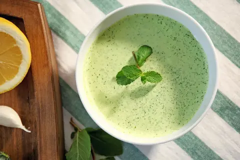 Un bol de sauce à la menthe fraîchement préparée, garni de feuilles de menthe. À côté, une tranche de citron et une gousse d'ail sur une planche en bois.