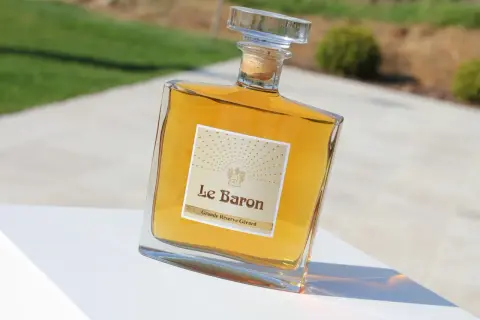 Bouteille de "Le Baron Grande Réserve Gérard", un spiritueux ambré, posée sur un socle blanc, avec un arrière-plan flou évoquant un jardin et un chemin, rappelant l'élégance d'un chateau vaudois.