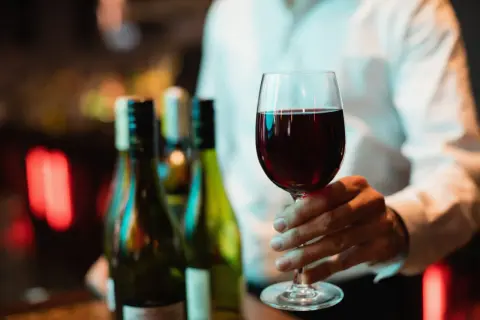 Main d'un homme tenant un verre de vin rouge, prêt pour une dégustation de vin Beaujolais. Des bouteilles floues en arrière-plan.