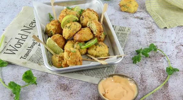 Plateau de délicieux accras dorés, garnis de quartiers de citron vert et de coriandre fraîche, servis avec une sauce crémeuse.