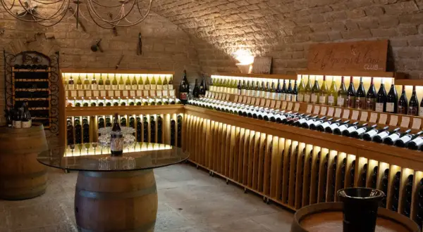 Cave voûtée du **domaine de Montmain** avec étagères en bois illuminées présentant de nombreuses bouteilles de vin, tonneaux et lustre ancien.