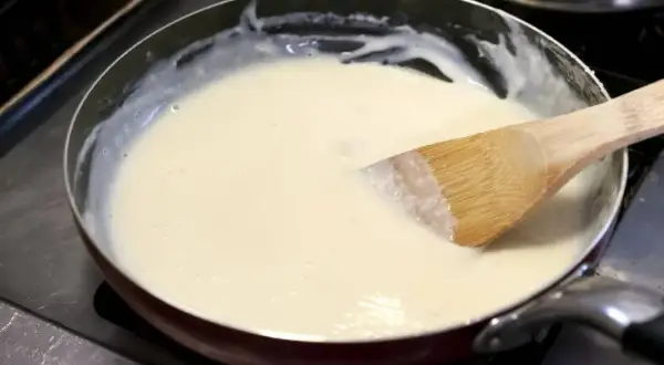 Une spatule en bois remue une sauce onctueuse dans une poêle. La préparation crémeuse, une sauce vin blanc et crème, est prête à être servie.