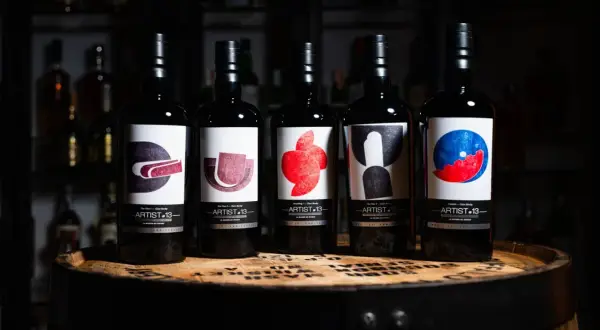 Cinq bouteilles de blend whisky Artist-13, ornées d'étiquettes artistiques colorées, reposent sur un fût de whisky.