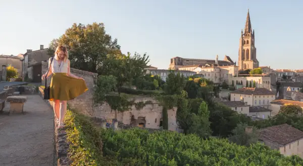 Une femme en jupe jaune marche sur un muret, surplombant le village de Saint-Émilion. La route des vins s'étend à perte de vue.