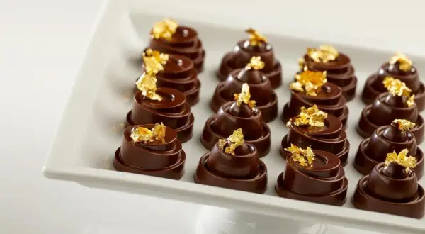Douze chocolats fins, une composition alimentaire luxueuse, ornés de feuilles d'or comestibles, présentés sur un plat blanc.