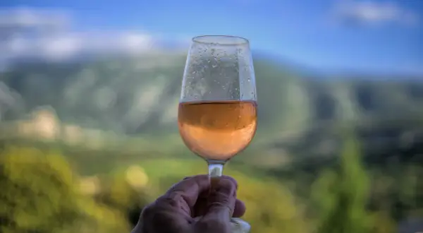 Un verre de vin rosé frais, avec des gouttes de condensation, tenu à la main devant un paysage de montagne flou. Un moment de détente avec un vin d'une région ensoleillée.