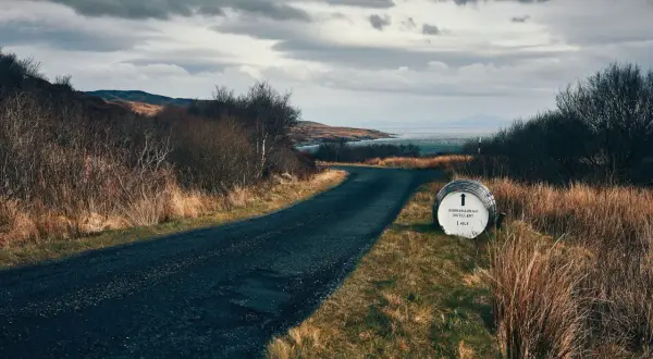 Route sinueuse menant à la distillerie Bunnahabhain, avec un fût indiquant la direction. Paysage écossais sauvage sous un ciel menaçant.