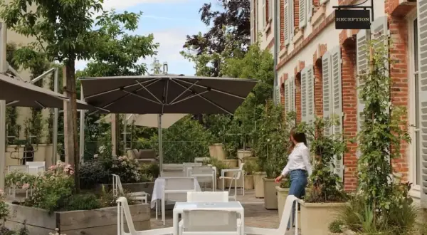 Terrasse verdoyante du 25bis by Leclerc Briant, avec tables, chaises et parasols. Une femme se dirige vers la réception.