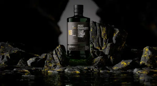 Bouteille de whisky Port Charlotte 18 ans d'âge, produit par la distillerie Bruichladdich, posée sur des rochers sombres aux reflets dorés.