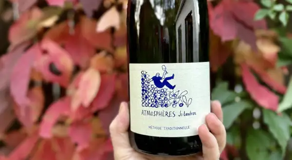 Une main tient une bouteille de vin nature Loire, "Atmosphères Jo Landron", devant des feuilles d'automne rouges et vertes.