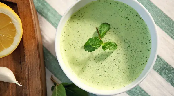 Un bol de sauce à la menthe fraîchement préparée, garni de feuilles de menthe. À côté, une tranche de citron et une gousse d'ail sur une planche en bois.