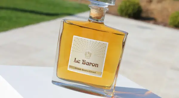 Bouteille de "Le Baron Grande Réserve Gérard", un spiritueux ambré, posée sur un socle blanc, avec un arrière-plan flou évoquant un jardin et un chemin, rappelant l'élégance d'un chateau vaudois.