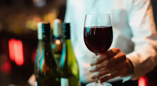 Main d'un homme tenant un verre de vin rouge, prêt pour une dégustation de vin Beaujolais. Des bouteilles floues en arrière-plan.