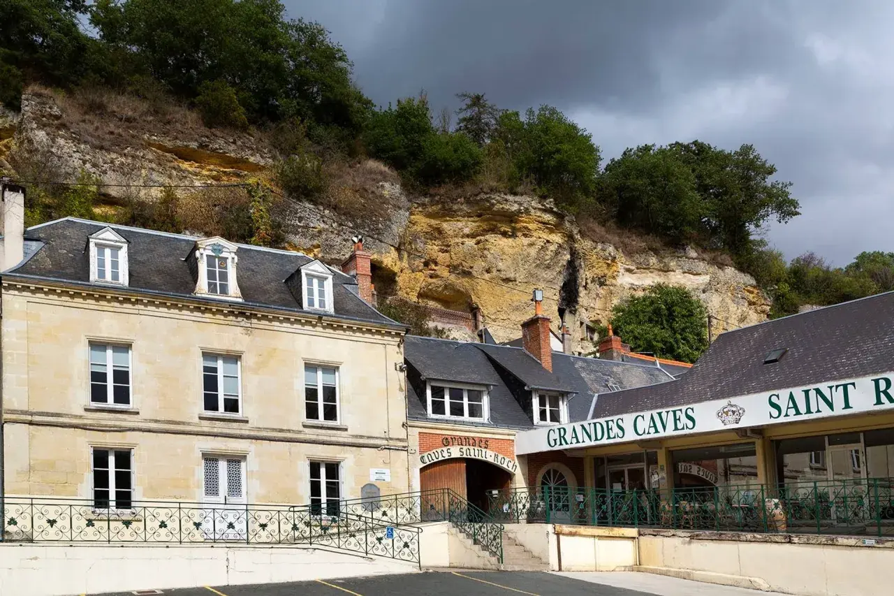 La maison Lacheteau, avec ses façades en pierre et ses toits d'ardoise, se dresse au pied d'une falaise rocheuse.