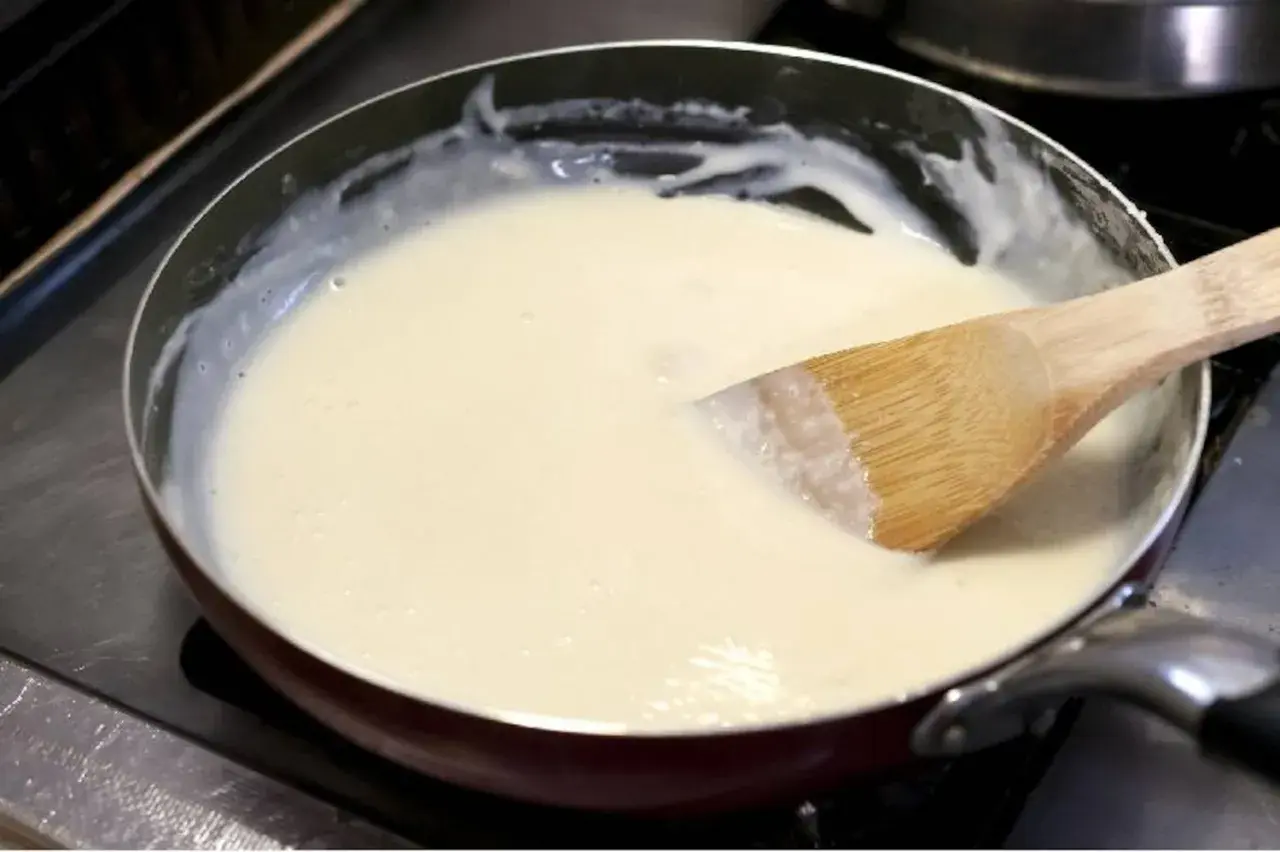 Une spatule en bois remue une sauce onctueuse dans une poêle. La préparation crémeuse, une sauce vin blanc et crème, est prête à être servie.