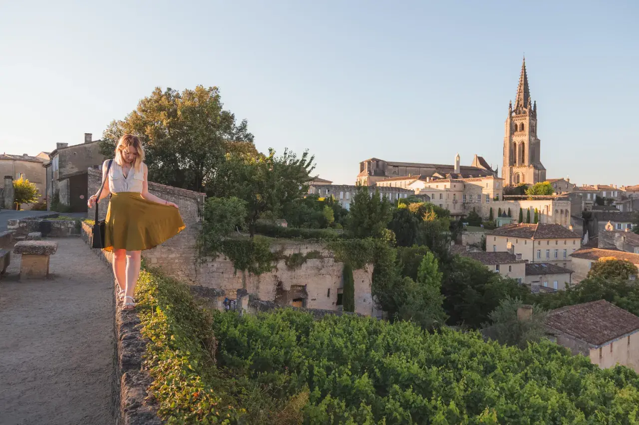 Une femme en jupe jaune marche sur un muret, surplombant le village de Saint-Émilion. La route des vins s'étend à perte de vue.