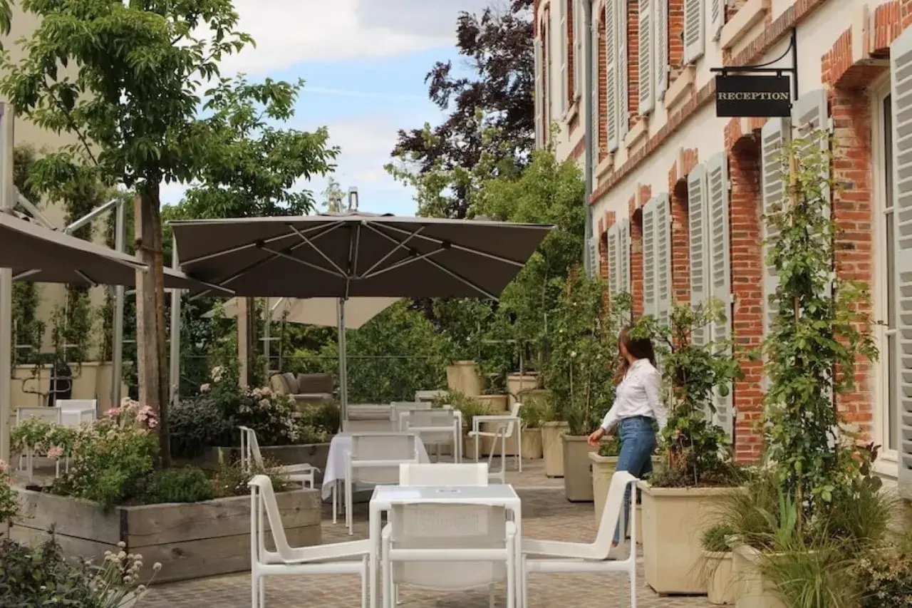 Terrasse verdoyante du 25bis by Leclerc Briant, avec tables, chaises et parasols. Une femme se dirige vers la réception.