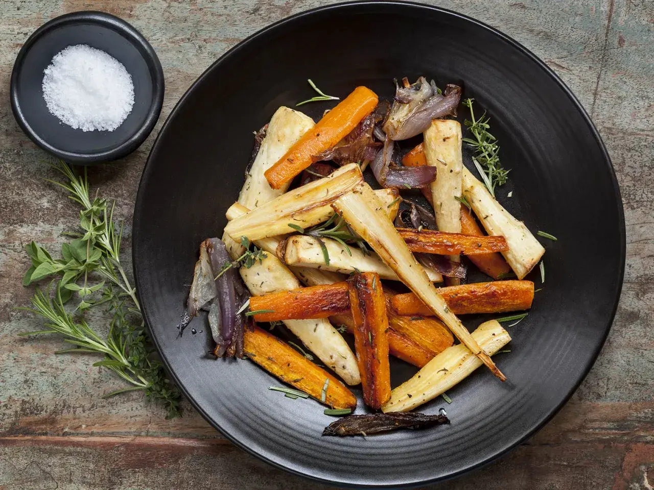 Légumes rôtis, carottes et panais, parsemés d'herbes. Un navet doré, tendre et savoureux, se mêle aux autres légumes sur l'assiette noire.