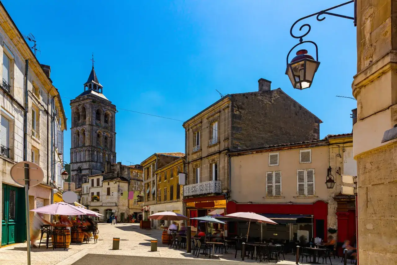 Une rue animée de la ville de Cognac, avec une tour d'église imposante en arrière-plan. Des terrasses de cafés invitent à la détente sous le soleil.