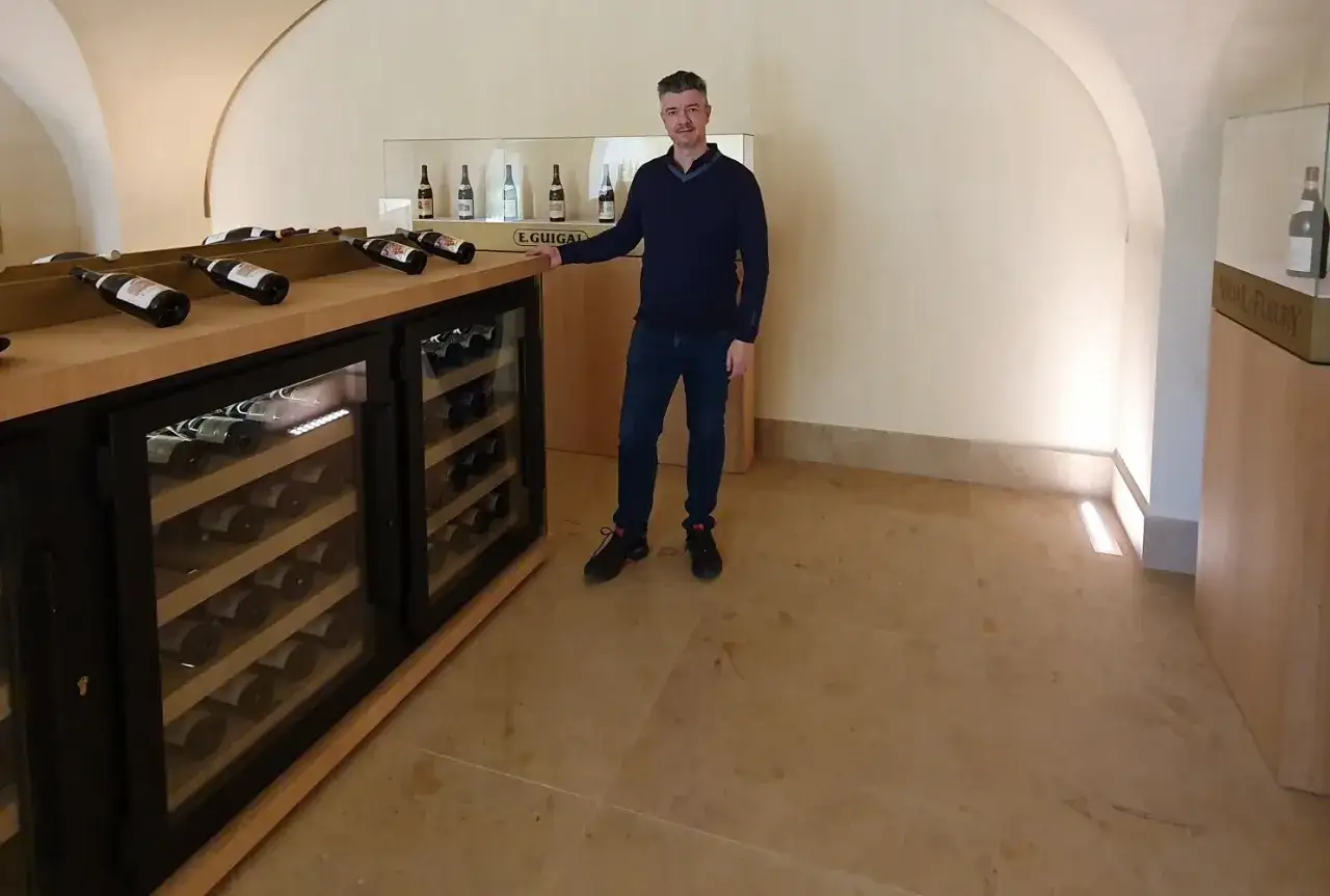 Homme souriant lors d'une visite cave à Ampuis, devant des réfrigérateurs à vin et des bouteilles exposées.