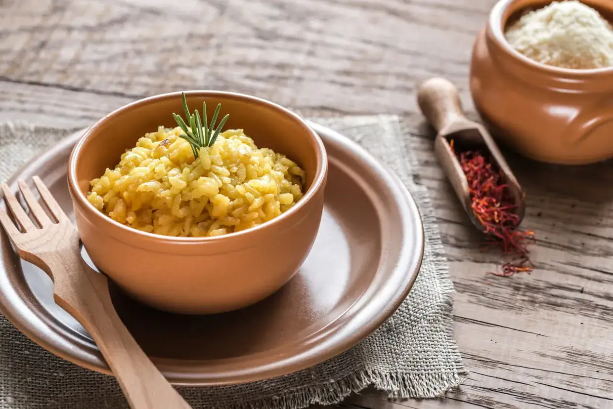 Un risotto dor&eacute;, parfum&eacute; au safran, est servi dans un bol en terre cuite. &Agrave; c&ocirc;t&eacute;, une cuill&egrave;re en bois r&eacute;v&egrave;le des pistils rouge intense.