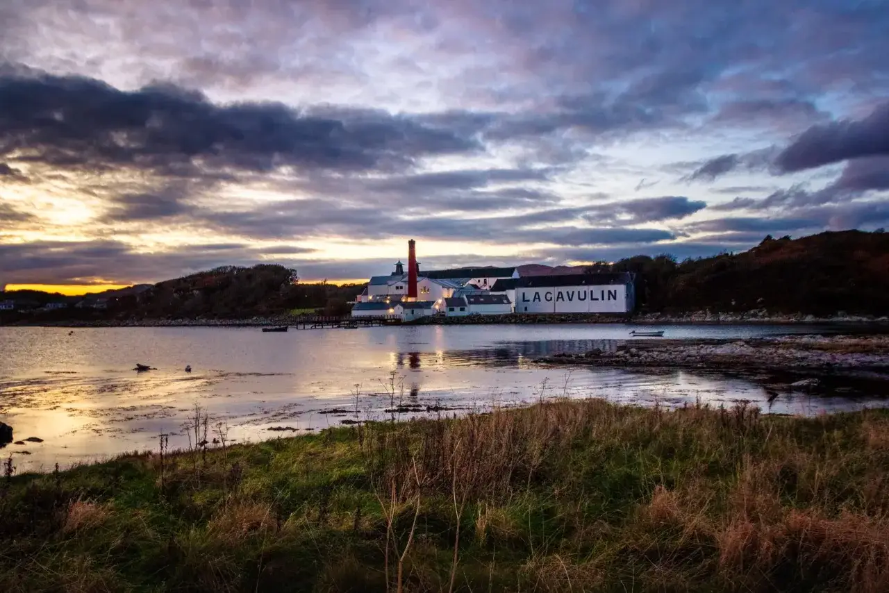 Coucher de soleil sur la distillerie Lagavulin, Islay. L'eau reflète le ciel dramatique, promettant un whisky tourbé authentique.