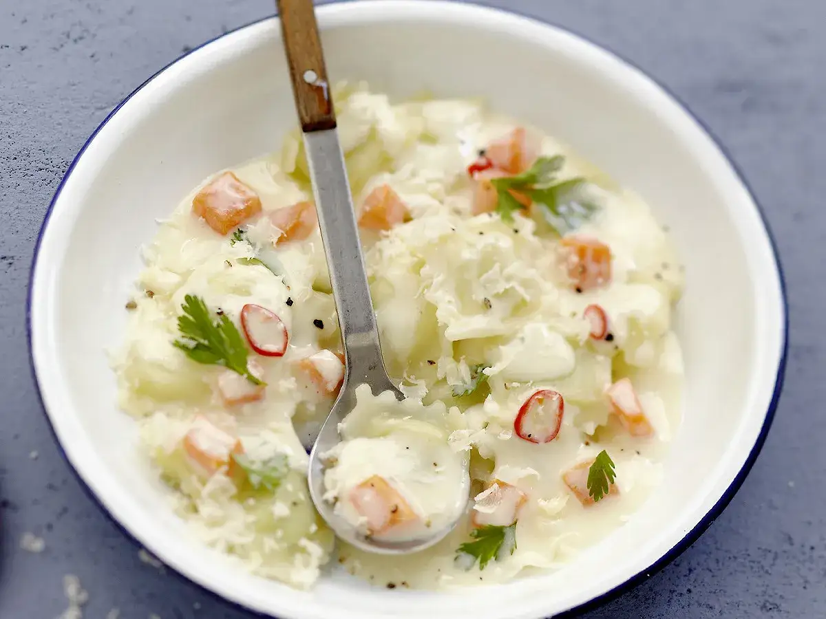 Un plat de ravioles de Royan crémeuses, parsemées de dés de carottes, de persil et de copeaux de parmesan. Une cuillère est plongée dans la sauce onctueuse.