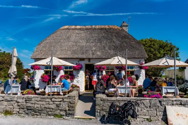 Café pittoresque avec toit de chaume, parasols et fleurs. Des gens dégustent un repas en plein air, peut-être un délicieux aran poisson.