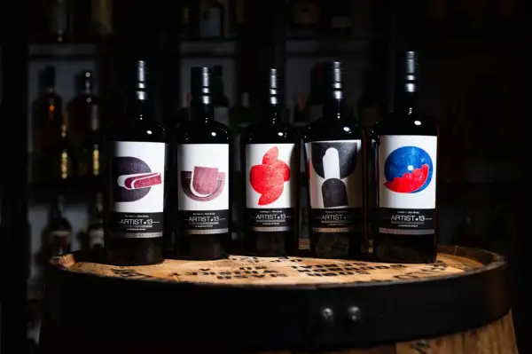 Cinq bouteilles de blend whisky Artist-13, ornées d'étiquettes artistiques colorées, reposent sur un fût de whisky.