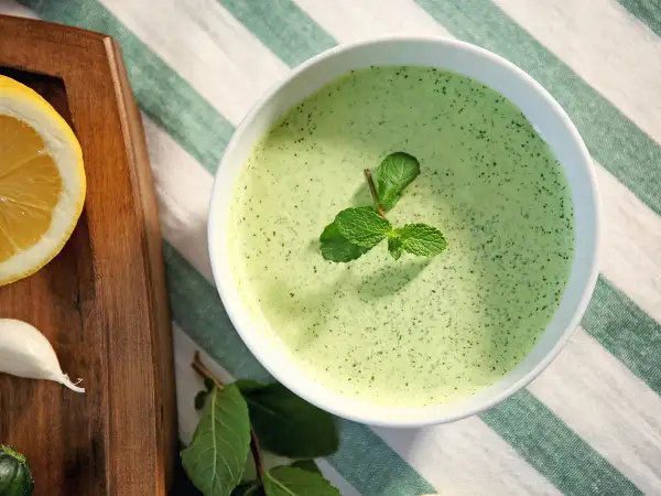 Un bol de sauce à la menthe fraîchement préparée, garni de feuilles de menthe. À côté, une tranche de citron et une gousse d'ail sur une planche en bois.