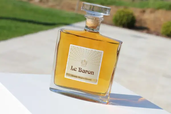 Bouteille de "Le Baron Grande Réserve Gérard", un spiritueux ambré, posée sur un socle blanc, avec un arrière-plan flou évoquant un jardin et un chemin, rappelant l'élégance d'un chateau vaudois.